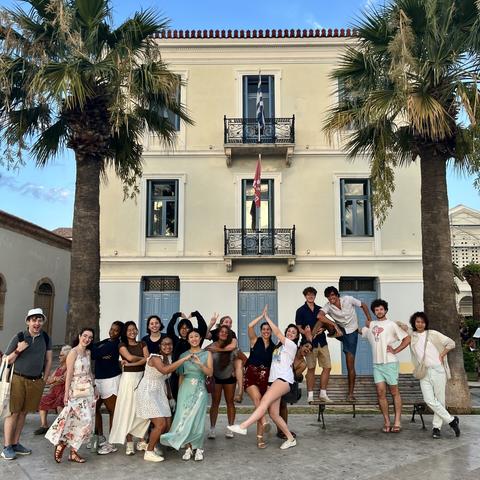 A group of young people pose joyfully in front of a neoclassical building with blue shutters, standing between two palm trees on a sunny day. Some are striking playful poses, and a few stand or sit on a bench.
