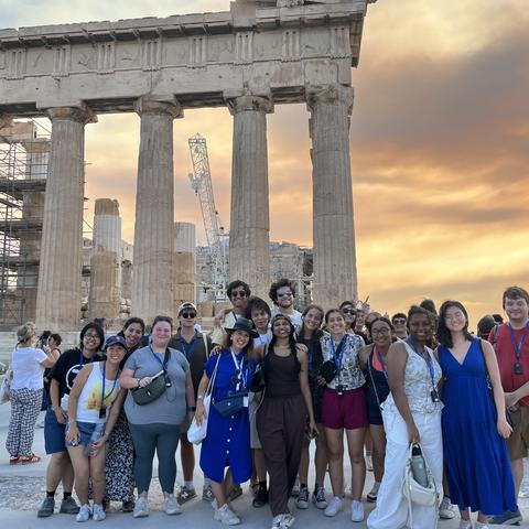 A group of people smile for a group photo in front of the ancient Greek Parthenon at sunset.
