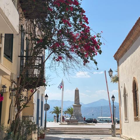 A sunlit street with a flowering bougainvillea, leading to a seaside promenade with a stone obelisk, palm trees, and mountains in the background.