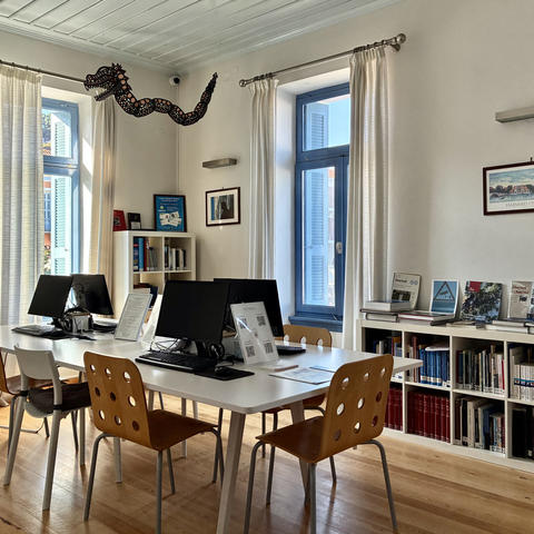 Bright study room with computers on a central table, bookshelves, and tall windows with light curtains.