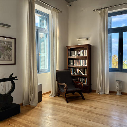 Quiet reading corner with a wooden armchair, bookcase, sculpture, and tall blue‑framed windows.