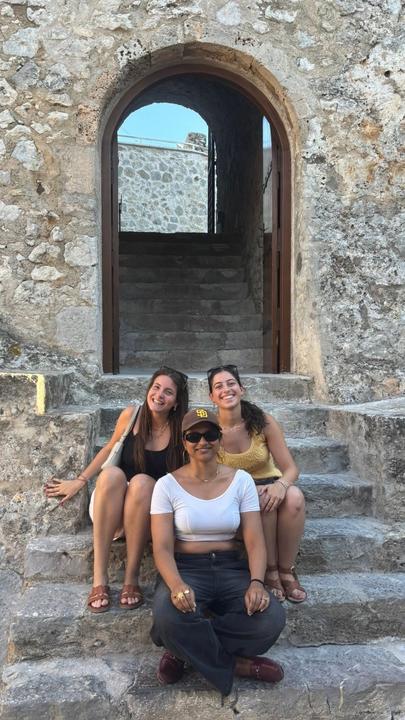Three young women sit on stone steps inside an arched doorway, all smiling and enjoying a sunny day.