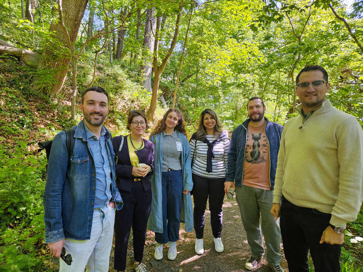 Six people standing and smiling on a sunlit wooded trail.