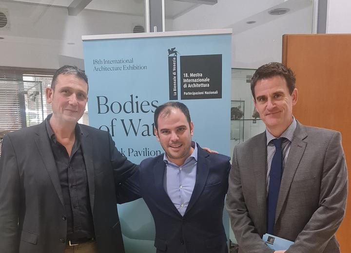 Three men standing and smiling at the camera in a room with a banner of the project Bodies of Water in the background.