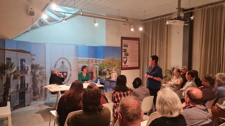 Inside a lecture hall, a member of the audience is standing up and speaking in front of two presenters and two dozens of listeners.