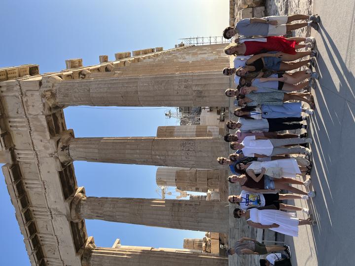 About 20 people posing for a group picture and smiling at the camera. Behind them, an ancient monument with tall columns can be seen.
