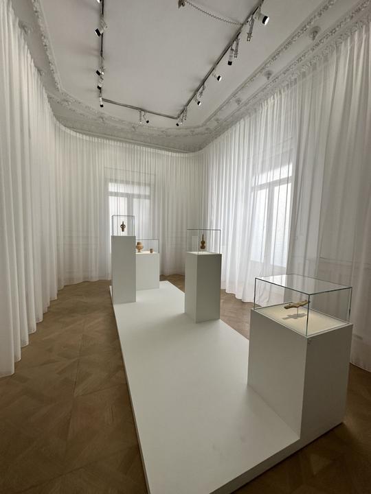 A room of elliptical design adorned with Cycladic figurines positioned prominently at its center. The windows are veiled with sheer, billowing white curtains, allowing a gentle diffusion of light.