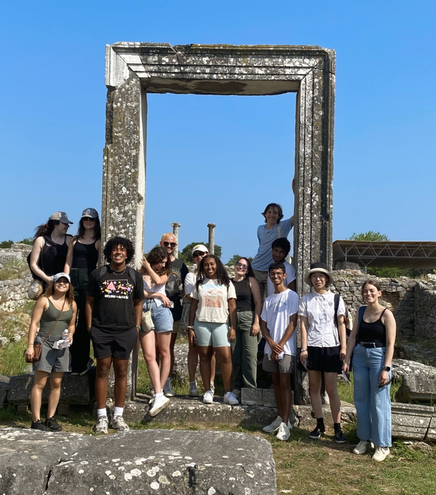 14 young people posing for a group picture and smiling at the camera. Ancient columns can be seen in the background while they are all scattered around an ancient rectangle-shaped column.