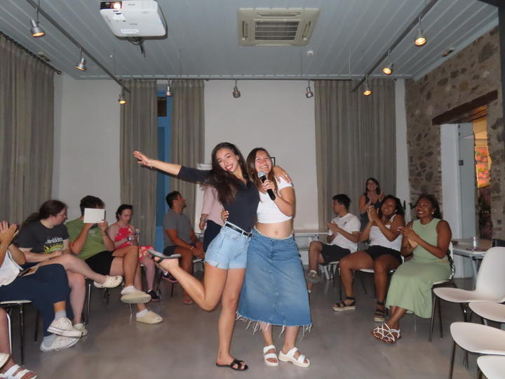 Two young women are standing and posing for the camera at the center of a room with curtains, stone walls, and overhead lighting. One is holding a microphone, while the other strikes a joyful pose with one leg raised. Around them, people are seated, clapping and smiling.