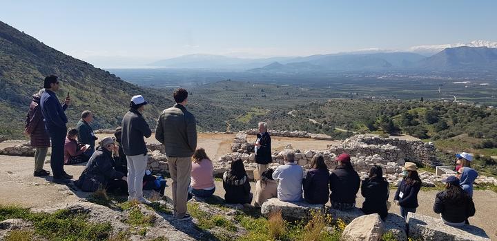 A group of twenty people seated between ancient monuments on a top of a hill and listening to one man standing in the middle.