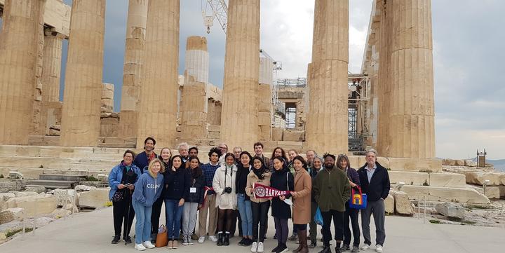 A group of twenty people facing the camera and holding a Harvard banner in front of the Parthenon of Athens.