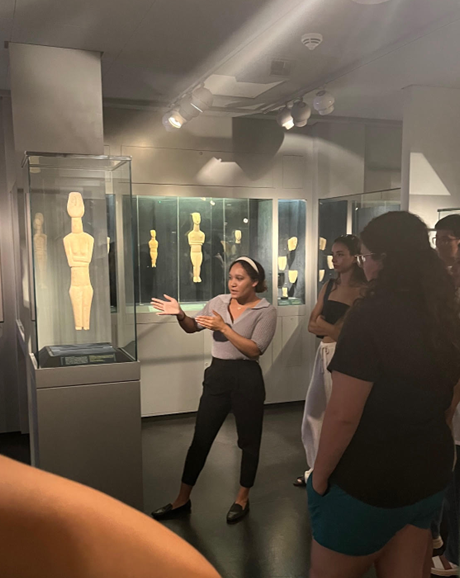 A young woman is standing in front of a glass display case containing a large, cycladic figurine. She is gesturing with her hands, explaining something to a small group of people gathered around her.