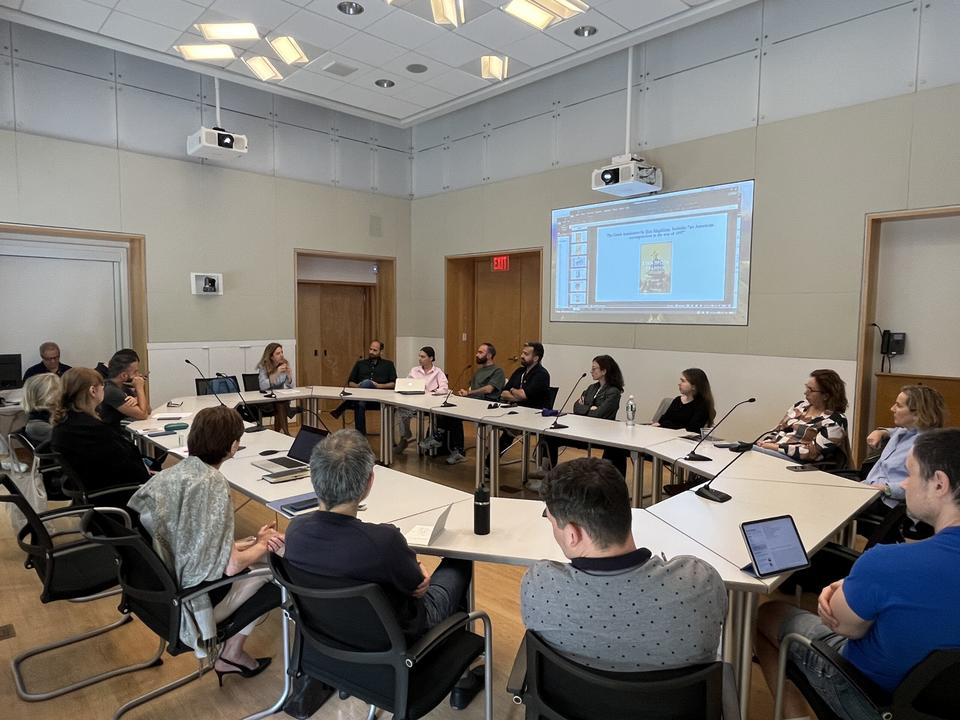 A group sits around a U-shaped table in a modern conference room, listening to a woman speak. Laptops and a projected slide are visible, indicating a formal meeting or seminar.