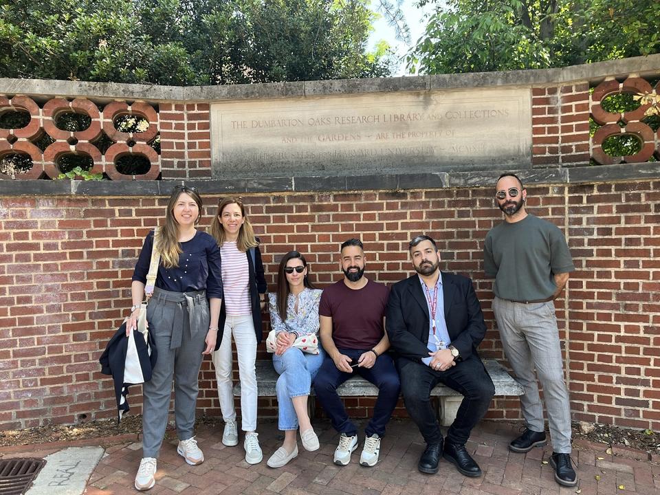 Six people pose in front of a brick wall with a plaque at Dumbarton Oaks, three sitting and three standing, with greenery above on a sunny day.