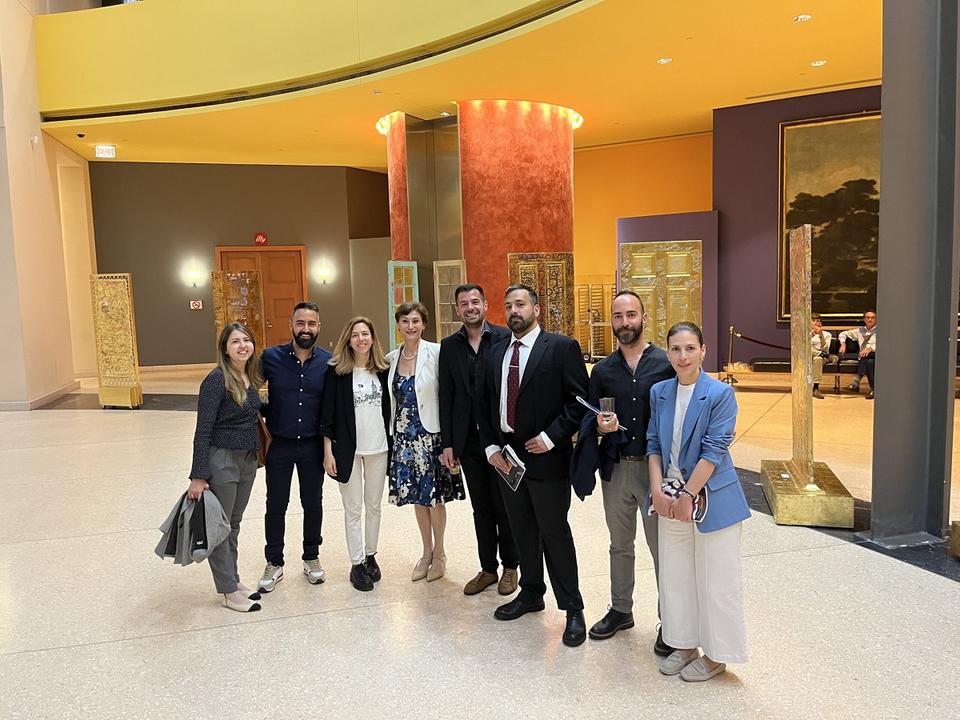 Eight people pose for a photo in a spacious, modern lobby with yellow and orange décor and ornate golden screens. They are dressed in smart casual and business attire, smiling in a friendly, professional setting.