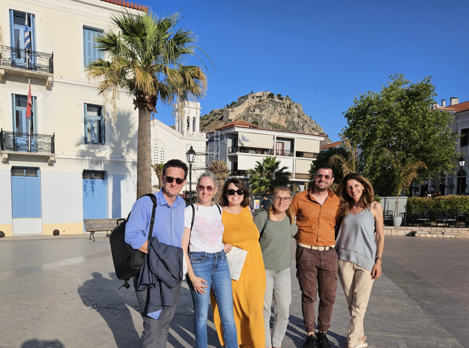 Six adults smile and pose together in a sunny town square. Behind them are a palm tree, pastel buildings, and a rocky hill with a fortress on top, all under a clear blue sky.