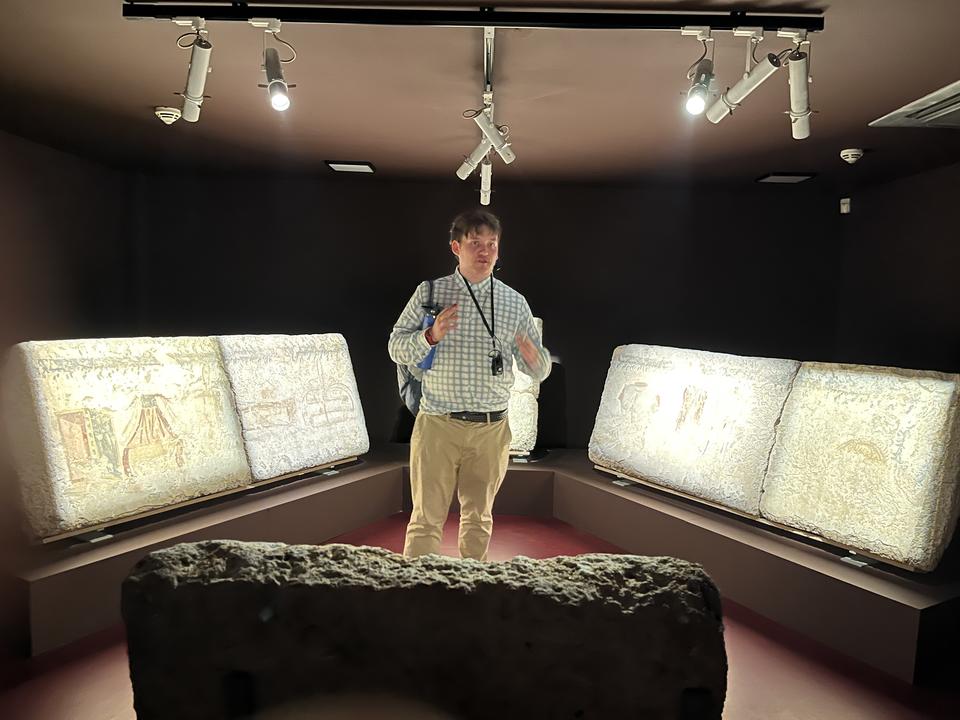A person stands in a dimly lit museum room, surrounded by illuminated ancient stone reliefs displayed on angled platforms along the walls. Overhead track lights highlight the artifacts, and the individual appears to be explaining or gesturing about the exhibits.
