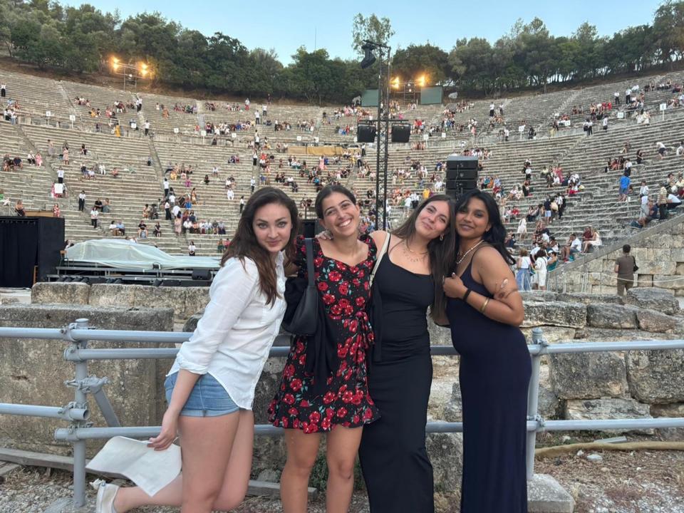 Four young women pose together in front of an ancient stone amphitheater, smiling at the camera, while people are dispersed across the large stone seating area in the background.