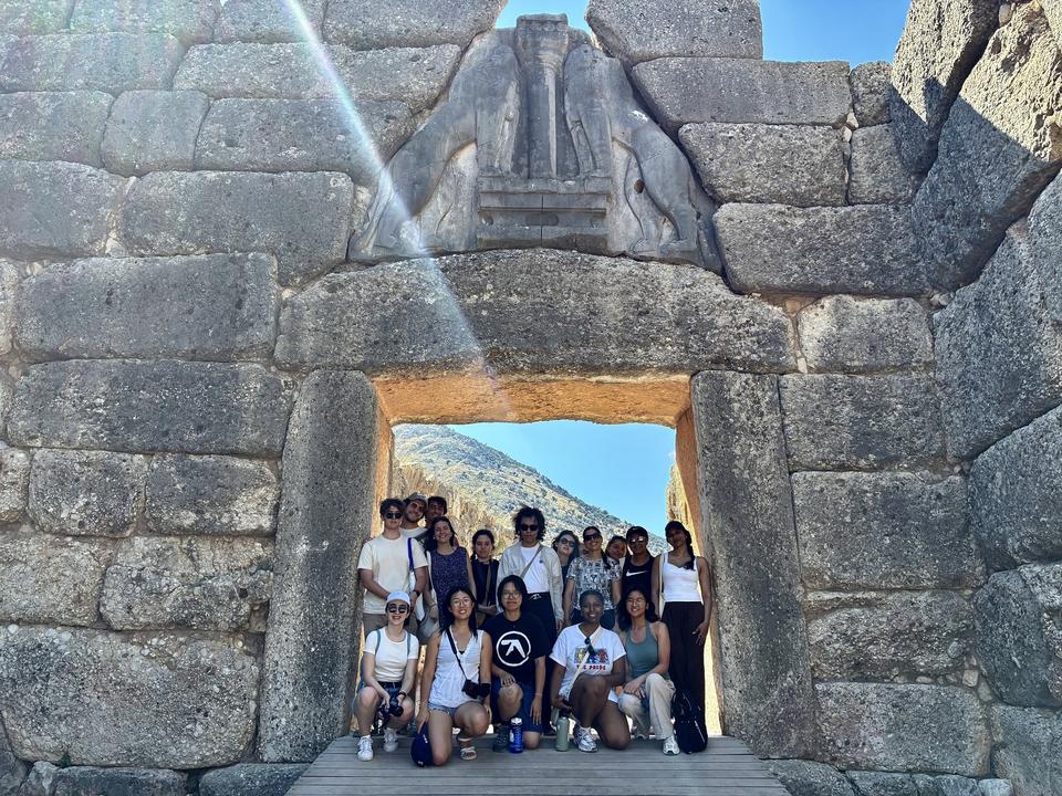 A group of people stand together for a photo beneath a massive ancient stone gate topped with carved lions, with sunlight shining over the scene and rugged mountains in the background.
