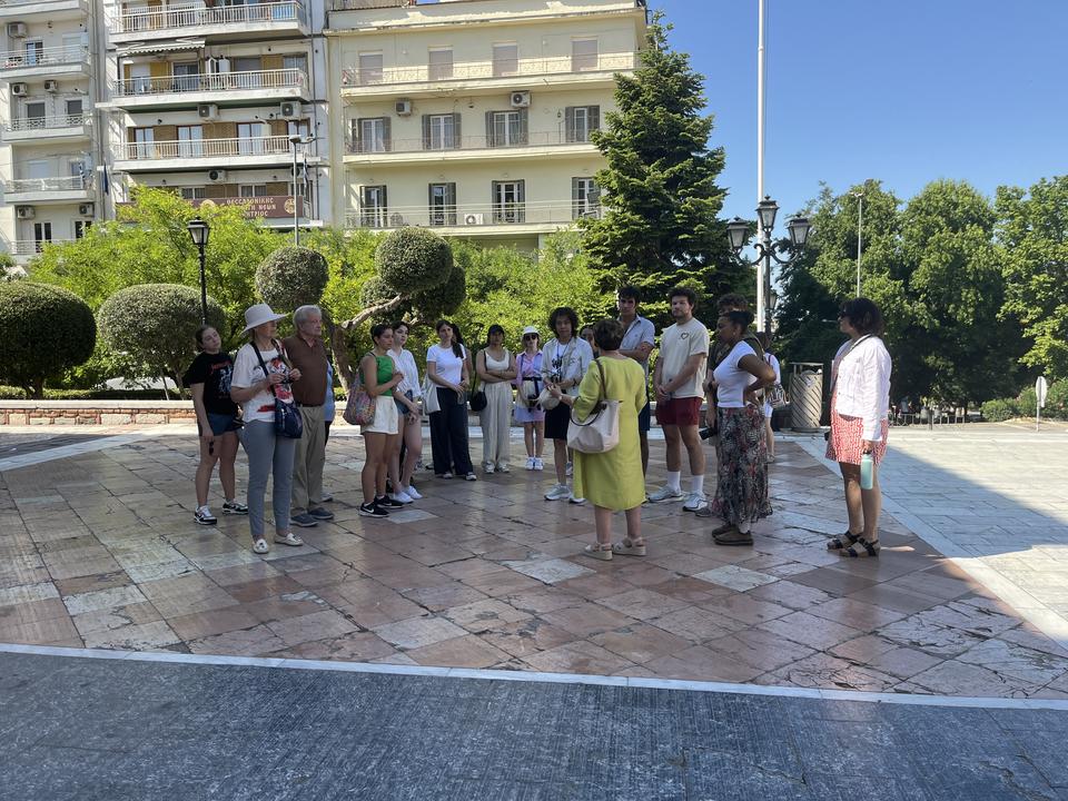 A group of people stands in a circle on a stone plaza surrounded by trees and apartment buildings, listening to a woman in a yellow dress speak to them.