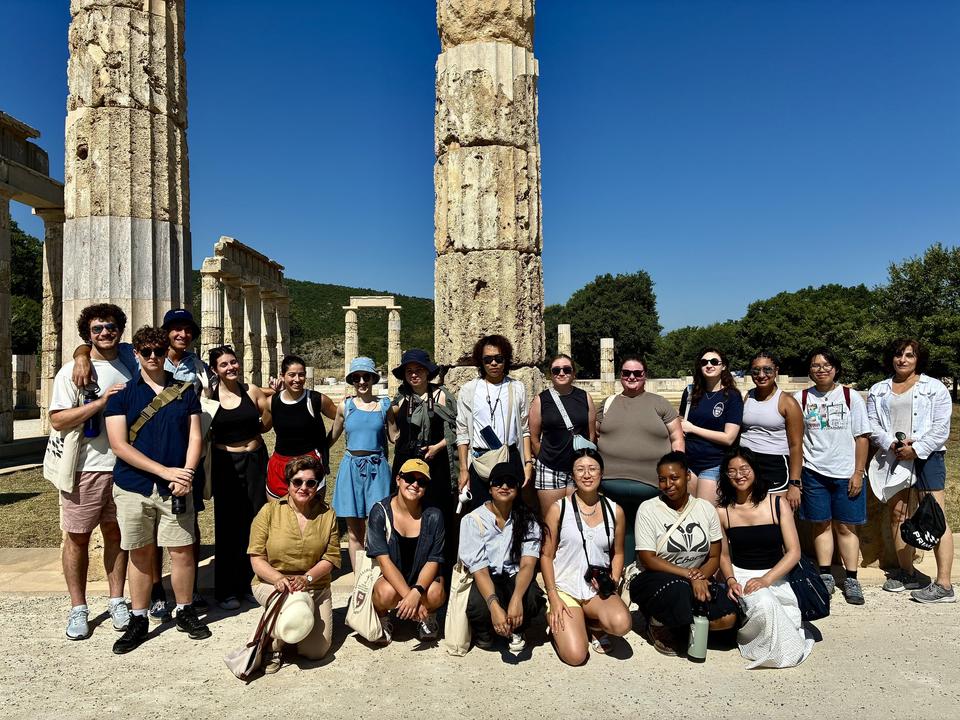 A large group of people, some standing and some sitting, take a group photo in front of tall, ancient stone columns and ruins under a bright blue sky.