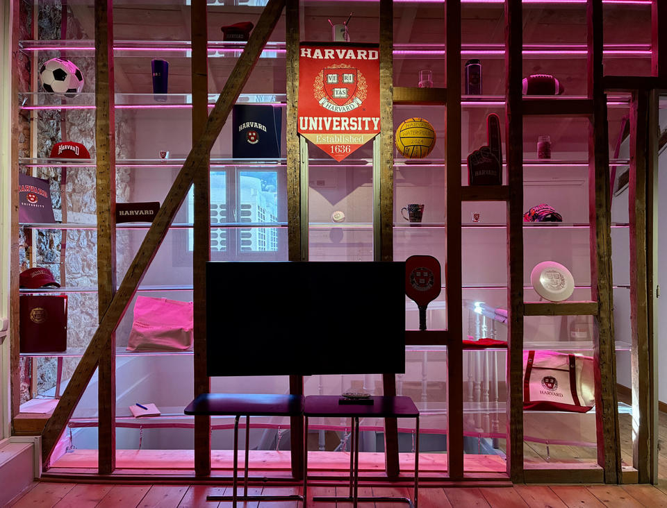 A display wall with wooden beams features Harvard memorabilia—such as hats, a soccer ball, mugs, a basketball, and a large Harvard banner—lit by pink lights. A screen on a stand sits in front of the display.