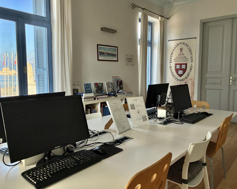 Bright study room with computers on a white table, wooden chairs, bookshelves, and large windows letting in sunlight.
