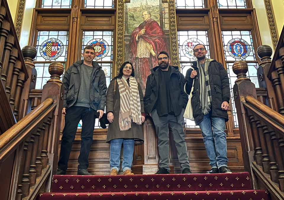 Four visitors standing on a grand wooden staircase in front of stained‑glass windows and a large painting of religious content.