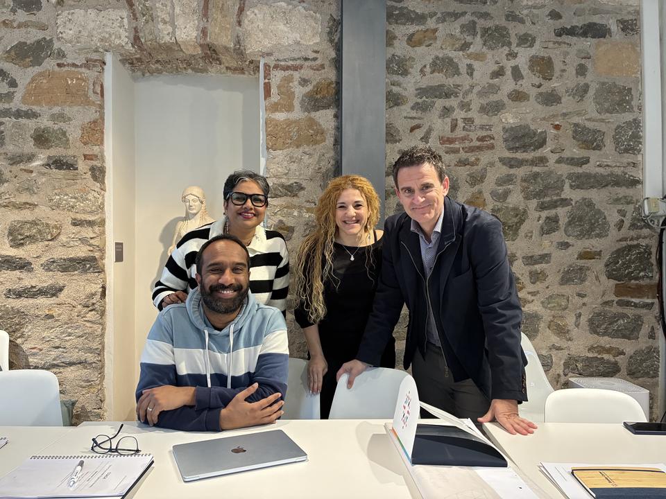 Four colleagues posing and smiling at a table in front of a stone wall, with notebooks and a laptop in front of them.