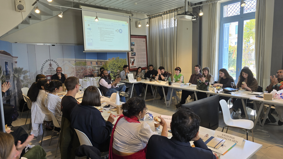 Large roundtable discussion in a bright room, with many attendees seated and facing a projected presentation on the wall.
