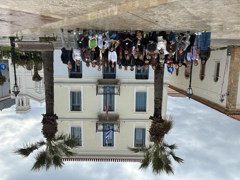 Large group photo of smiling people standing and kneeling in front of a neoclassical building with two palm trees.
