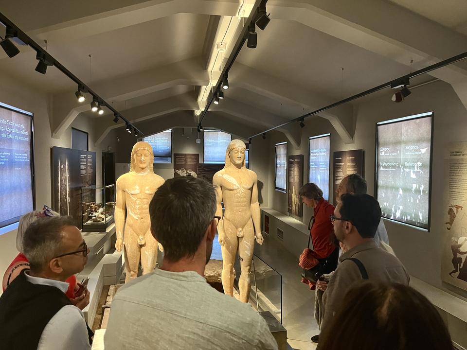 Visitors in a museum gathered around two large ancient Greek kouros statues.