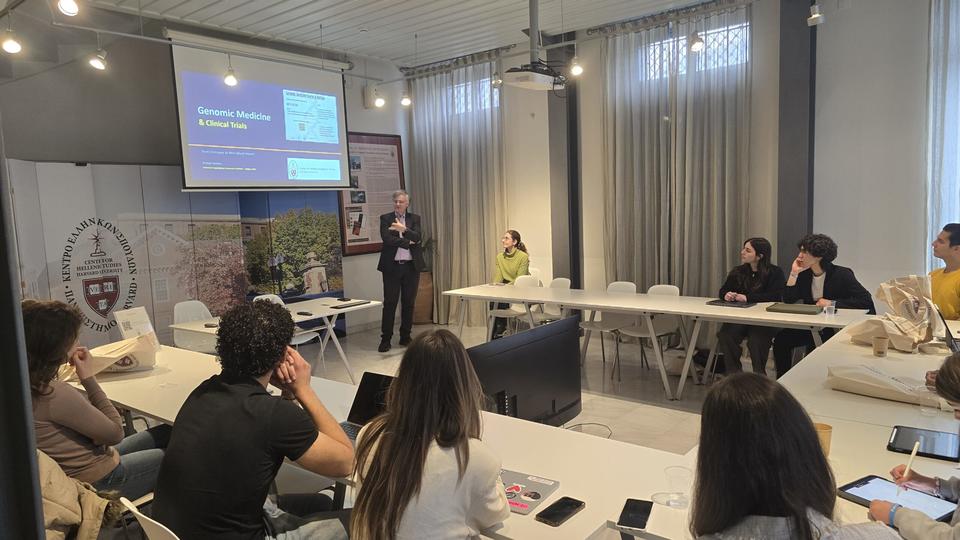 Lecture on genomic medicine and clinical trials in progress at CHS Greece, with students seated around tables and slides projected on the screen.
