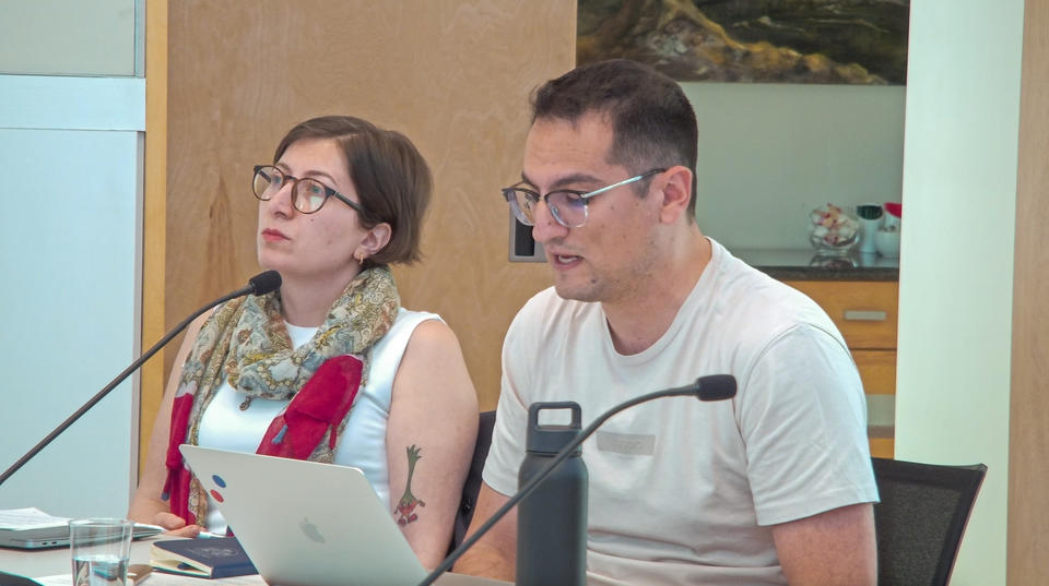 Two people seated at a table with microphones, with one of them speaking, in a conference room.
