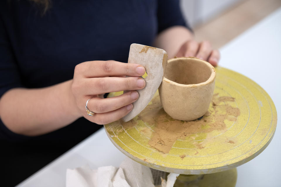 Person shaping a small clay cup on a pottery wheel with a smoothing tool.