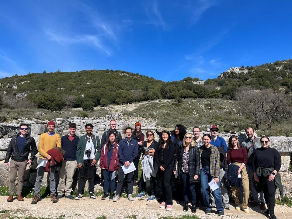 18 individuals pose for a group picture on a sunny day in front of ancient walls and a hilltop.