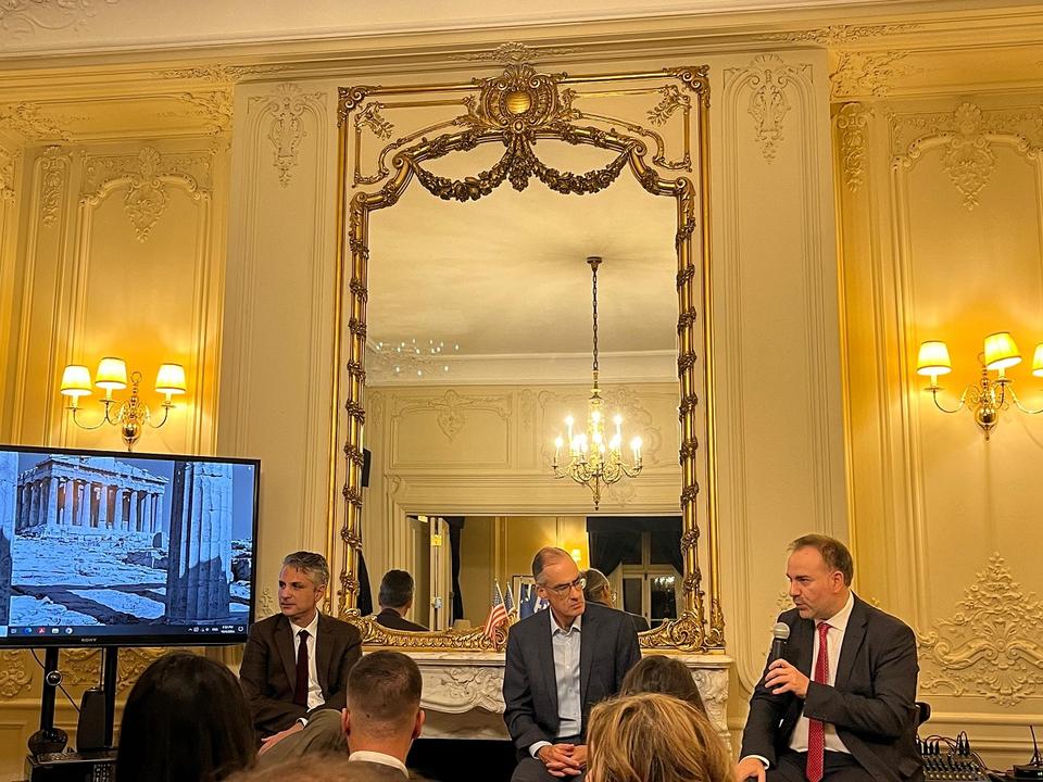 Three men seated in an elegant, ornately decorated room, engaged in a discussion. The man on the right holds a microphone, speaking to the audience, while the other two listen attentively. Behind them, a large, gilded mirror reflects a chandelier and intricate wall moldings, while a screen on the left displays an image of the Parthenon.