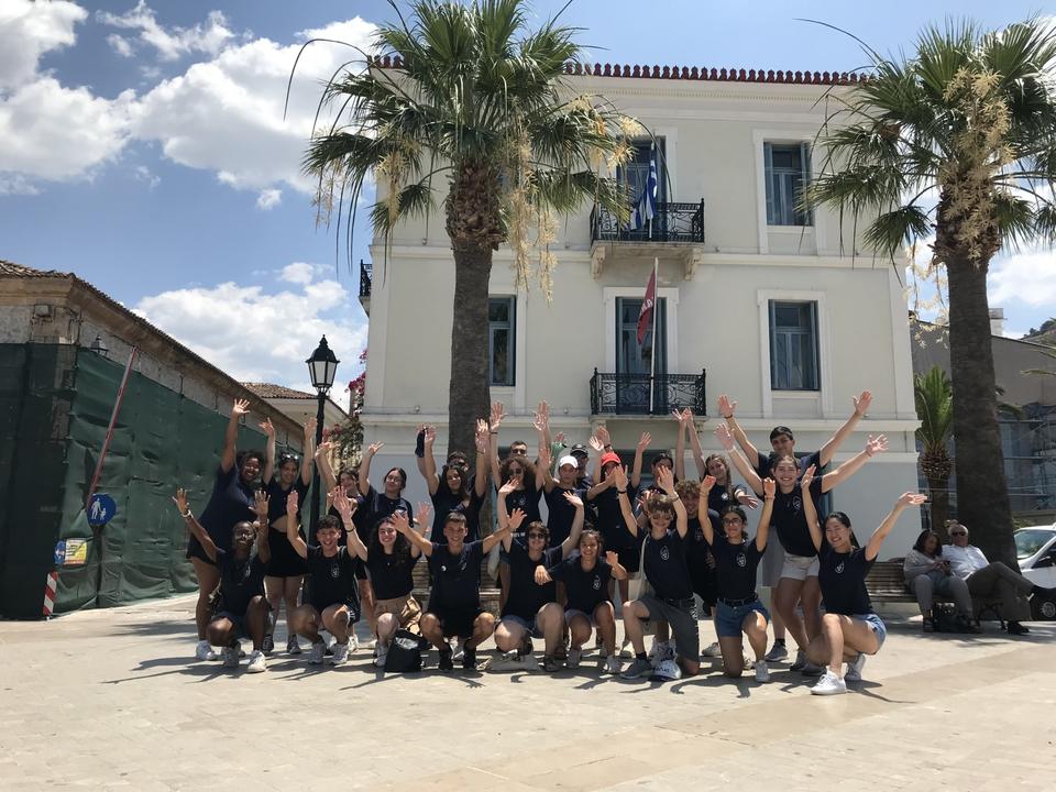 A group of 25 young people standing in front of a white building, all wearing the same T-shirts, with their hands raised and smiling.