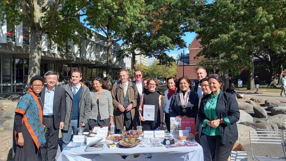 A smiling and diverse group of individuals standing outdoors beside an information table, which is covered with informational materials, pamphlets, candy bowls, and signs encouraging to stay updated with Harvard's global outreach.