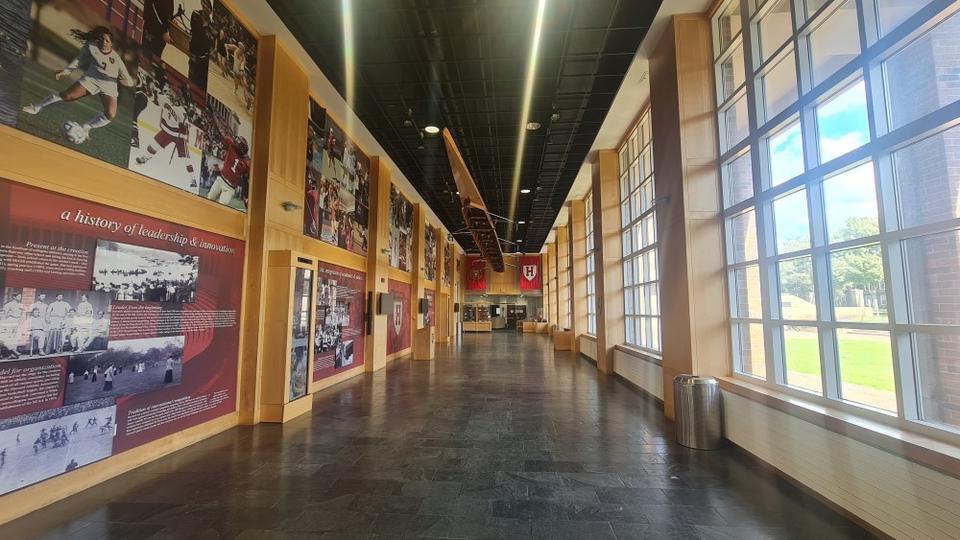 A spacious corridor with windows on the right and a wall with pictures of athletes from various sports on the left. The message 'a history of leadership & innovation' along with additional informative details are displayed. A canoe is suspended from the ceiling and, at the end of the hallway, Harvard logo banners are on display.