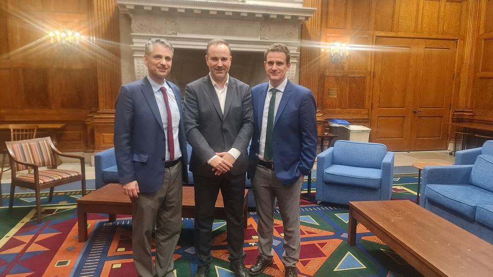 Three smiling men standing side by side in a formal setting, each dressed in suits, in front of a large stone fireplace with intricate carvings. The room has wooden paneling, blue upholstered chairs, and a colorful geometric-patterned rug.