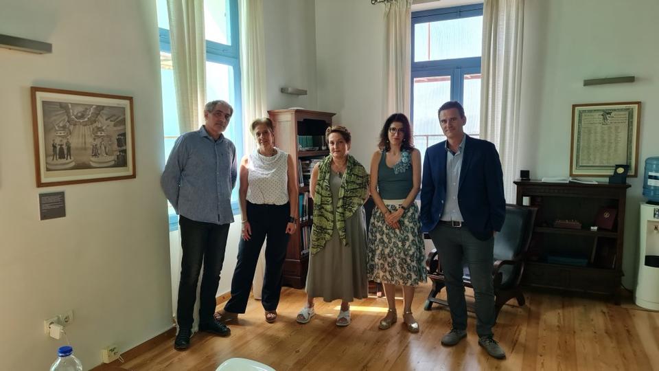Five people, including two men on the sides and three women in the middle, stand in a room with a wooden floor. They are positioned between sun-lit windows and are looking directly at the camera.
