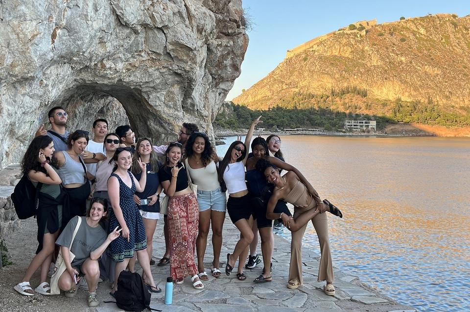 A group of people is posing for the camera in a friendly manner, with some striking joyful poses and one gesturing the victory sign. They are standing on a pathway by the sea, with a large rocky passage as their backdrop on the left side and a sunlit hill on the right.