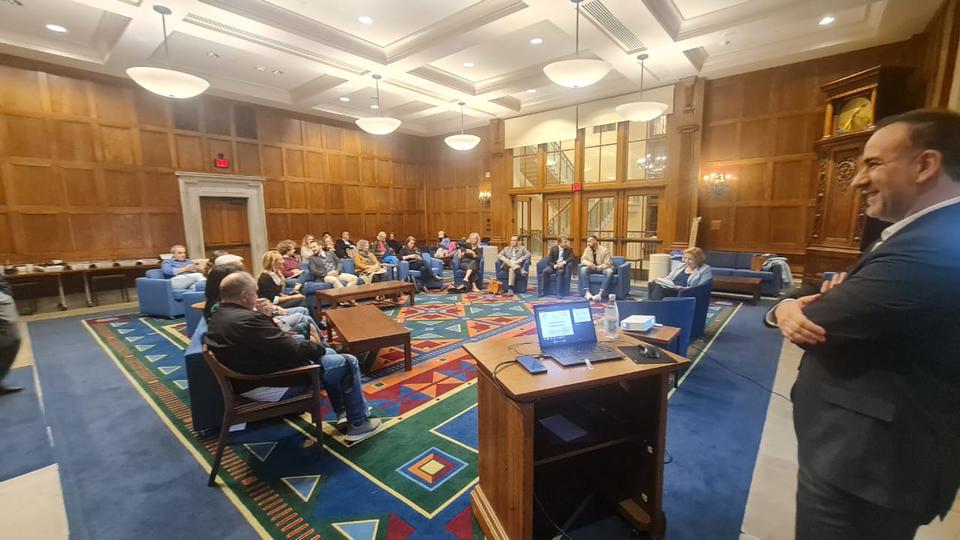 A group of people is seated in a semi-circle at a large, wood-paneled room with a high ceiling and warm lighting. In the foreground, a man stands to the right, smiling, with a laptop set up on a wooden podium displaying a presentation slide. The floor is covered with a colorful geometric-patterned rug. The setting suggests a formal yet comfortable discussion.