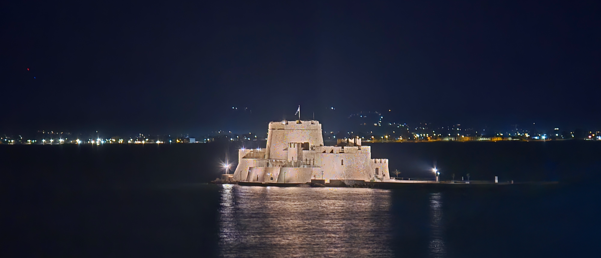 Stone fortress on a small island illuminated at night, surrounded by calm dark water and distant city lights on the horizon.