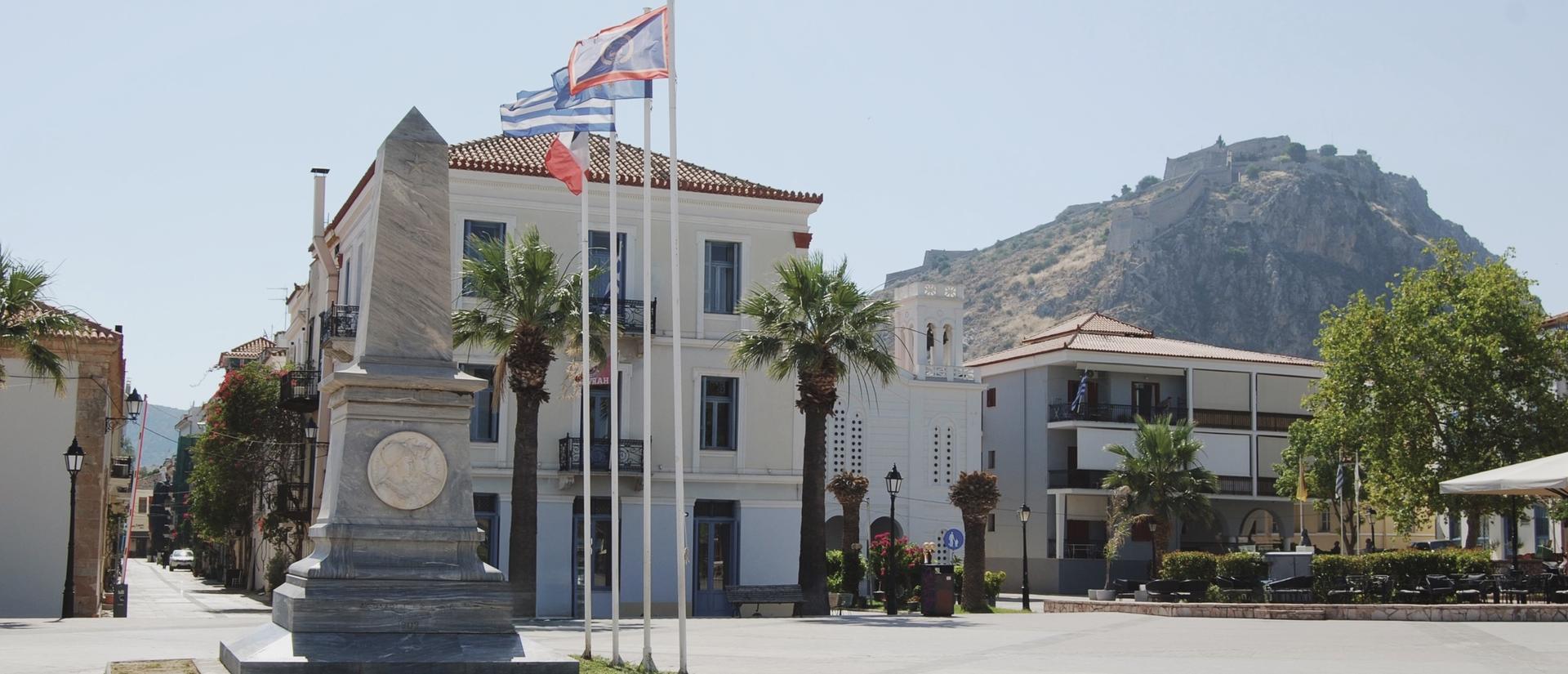 In the foreground, a marble monument stands in a sunlit town square. Behind it rises the cream-colored CHS Greece building with blue window frames and palm trees, while the Palamidi Fortress crowns the hillside in the background beneath a clear blue sky.
