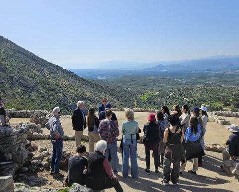 A group stands and sits among ancient stone structures on a hillside, attentively listening to a woman talking.