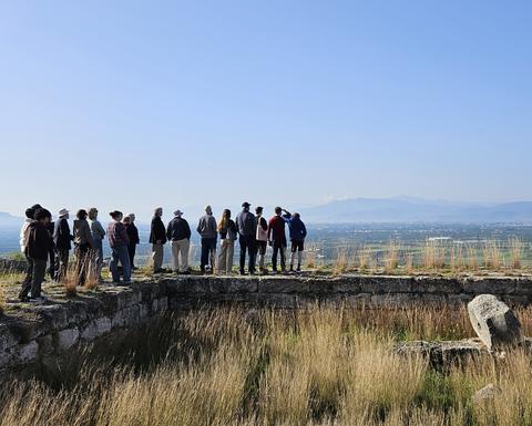 A group of people is gathered on the remains of an ancient stone structure, gazing out over a wide valley with scattered towns and distant mountains beneath a clear blue sky.
