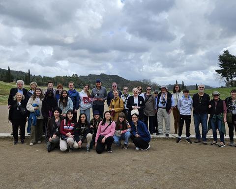 A large group of people, of various ages, pose for a group photo on a wide, open ancient stadium. 