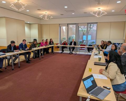Participants seated around U-shaped tables during a meeting in a conference room with laptops, documents, and large windows.